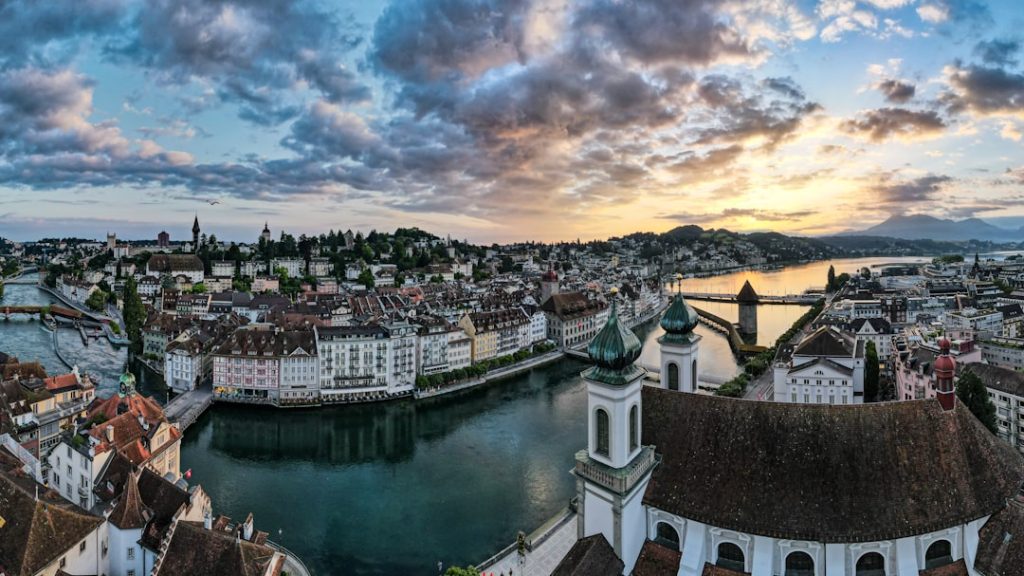 This sweeping panoramic view captures the waking city of Lucerne from an elevated vantage point. In the foreground, the iconic green domes of the Jesuit Church stand tall, overlooking the turquoise waters of the Reuss River as it flows toward the historic Chapel Bridge. The early morning sky is a canvas of dramatic, textured clouds, painted with the soft transitions of dawn—from deep cool blues to a warm, sun-drenched horizon. This composition highlights the medieval charm and architectural grandeur of Switzerland, offering a serene, storytelling perspective on a timeless European gem.